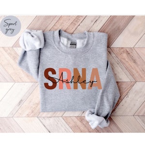 May include: A gray sweatshirt with the letters "SRNA" in brown, pink, and orange, with the name "Ashley" written in black cursive underneath.