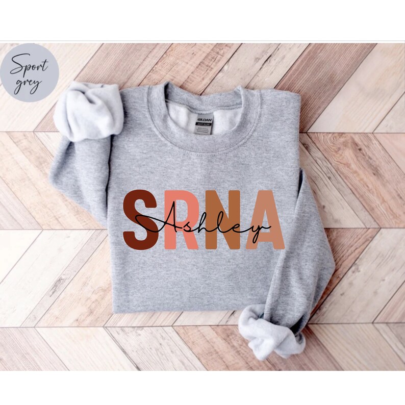 May include: A gray sweatshirt with the letters "SRNA" in brown, pink, and orange, with the name "Ashley" written in black cursive underneath.