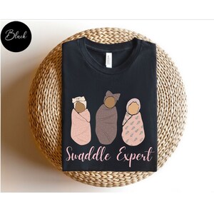 May include: Black t-shirt featuring a graphic of three babies swaddled in different patterns. The text "Swaddle Expert" is printed below the babies.