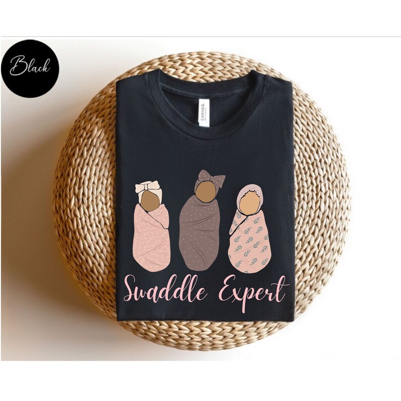 May include: Black t-shirt featuring a graphic of three babies swaddled in different patterns. The text "Swaddle Expert" is printed below the babies.