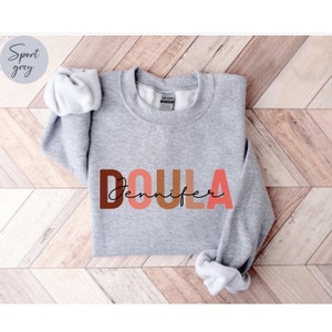 Pu&ograve; includere: Una felpa grigia con la parola "DOULA" in lettere colorate e il nome "Jennifer" scritto in corsivo nero sotto.