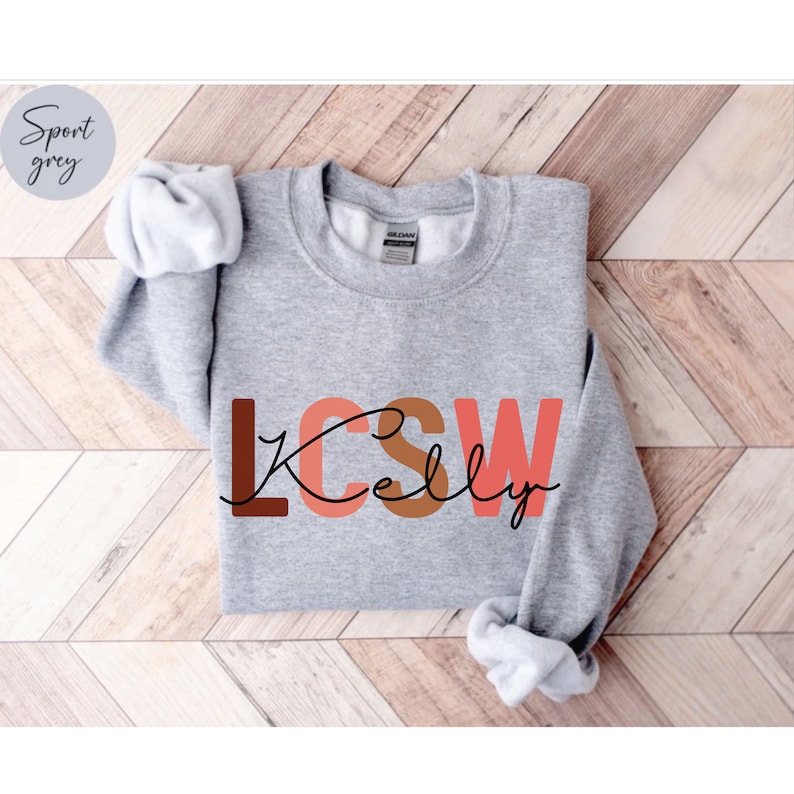 K&ouml;nnte beinhalten: Ein grauer Pullover mit dem Text "LCSW Kelly" in braunen, rosa und orangen Buchstaben. Die Buchstaben sind schwarz umrandet.