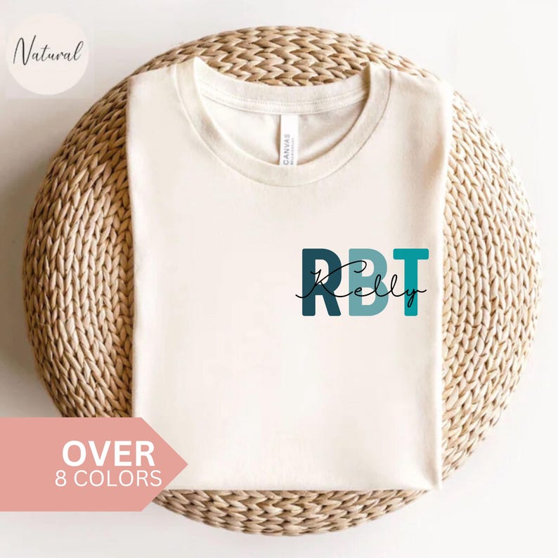 May include: Cream-colored t-shirt with the text "RBT Kelly" in teal and dark teal. The shirt is displayed on a woven, circular surface. The words "Over 8 Colors" are in a pink banner.