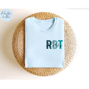 May include: A light blue t-shirt with the text "RBT Kelly" printed on the front in dark blue and teal. The shirt is displayed on a woven, circular surface. The upper left corner of the image has a light blue circle with the words "Heather ice blue".