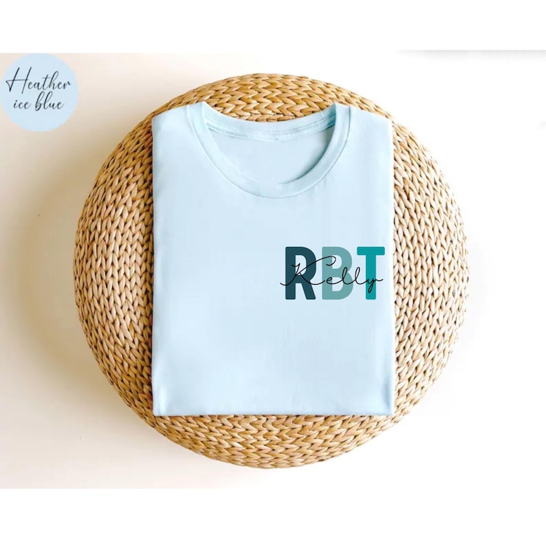 May include: A light blue t-shirt with the text "RBT Kelly" printed on the front in dark blue and teal. The shirt is displayed on a woven, circular surface. The upper left corner of the image has a light blue circle with the words "Heather ice blue".