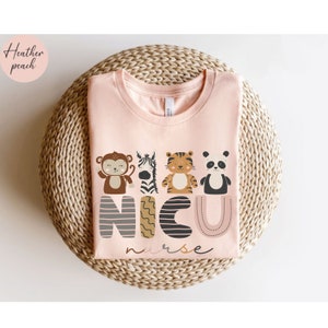 Pode incluir: Uma t-shirt rosa claro com um design de animais de desenho animado e o texto "NICU nurse". Os animais incluem um macaco, uma zebra, um tigre e um panda. A t-shirt &eacute; exibida numa superf&iacute;cie circular tecida.