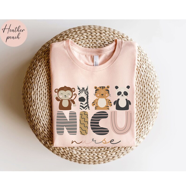 Pode incluir: Uma t-shirt rosa claro com um design de animais de desenho animado e o texto "NICU nurse". Os animais incluem um macaco, uma zebra, um tigre e um panda. A t-shirt &eacute; exibida numa superf&iacute;cie circular tecida.