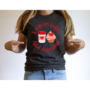 K&ouml;nnte beinhalten: Ein dunkelgraues T-Shirt mit dem Text "I run on coffee and cupcakes" in roten Buchstaben. Auf dem Shirt sind ein Cartoon-Kaffeebecher und ein Cupcake mit Herzen.