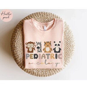 May include: A pink t-shirt with the words "PEDIATRIC ONCOLOGY" in a colorful, playful font. The letters are decorated with animal illustrations, including a monkey, zebra, tiger, and panda.