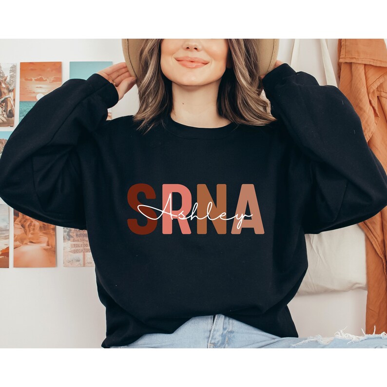 May include: Black sweatshirt with the text "SRNA" in colorful block letters and the name "Ashley" written in white cursive underneath.