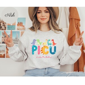 以下が含まれることがあります： カラフルな恐竜が「PICU nurse」と綴られているグレーのトレーナーです。