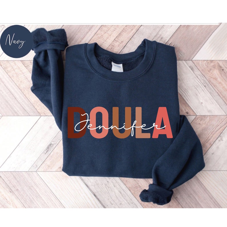 Pu&ograve; includere: Felpa blu navy con la parola "DOULA" in lettere maiuscole colorate e il nome "Jennifer" scritto in corsivo bianco.