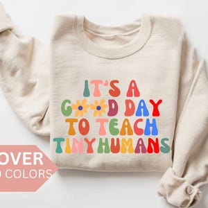 Op de afbeelding: Een beige sweatshirt met de tekst "It's a good day to teach tiny humans" in regenboogkleuren. De tekst is in een retro-lettertype met bloemen tussen de woorden "good" en "day".