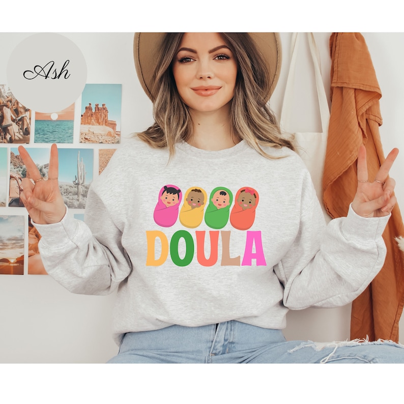 May include: A grey sweatshirt with a graphic of four babies wrapped in colorful blankets and the word "DOULA" in rainbow letters.