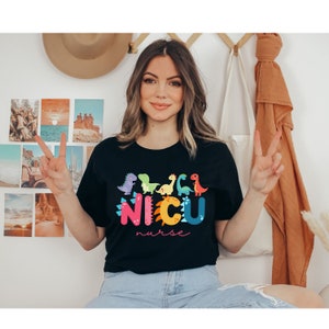 Pu&ograve; includere: T-shirt nera con dinosauri a fumetti colorati e la scritta "NICU nurse". I dinosauri sono in tonalit&agrave; di viola, verde, giallo e rosso. La parola "NICU" &egrave; in rosa, arancione e blu.