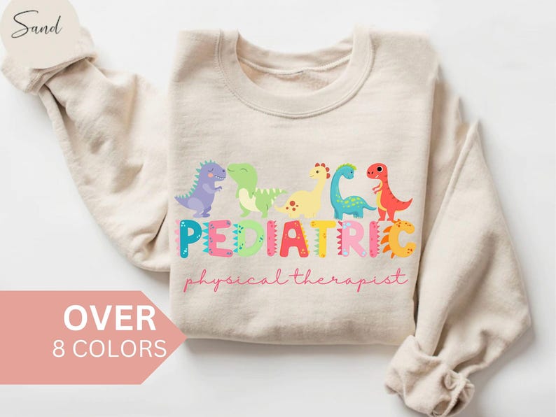 May include: A beige sweatshirt with a graphic design of five colorful dinosaurs spelling out the word 'PEDIATRIC' with the words 'physical therapist' below. The sweatshirt is available in over eight colors.