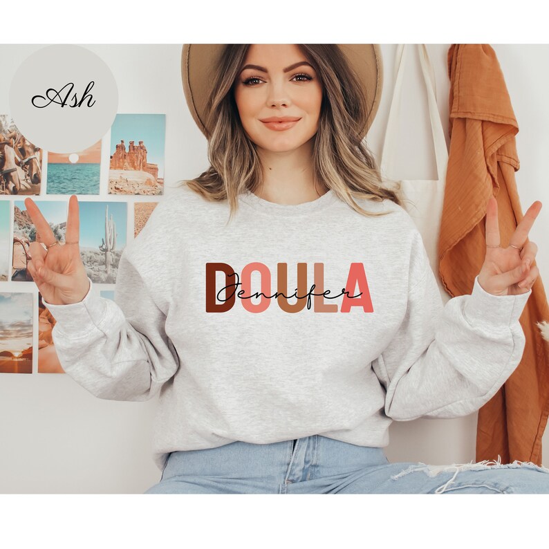 Pu&ograve; includere: Una felpa grigia con la parola "DOULA" stampata in lettere marroni, rosa e arancioni. Il nome "Jennifer" &egrave; scritto in corsivo nero sotto la parola "DOULA".