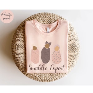 May include: A pink t-shirt with the text "Swaddle Expert" and an illustration of three babies swaddled in different patterns.