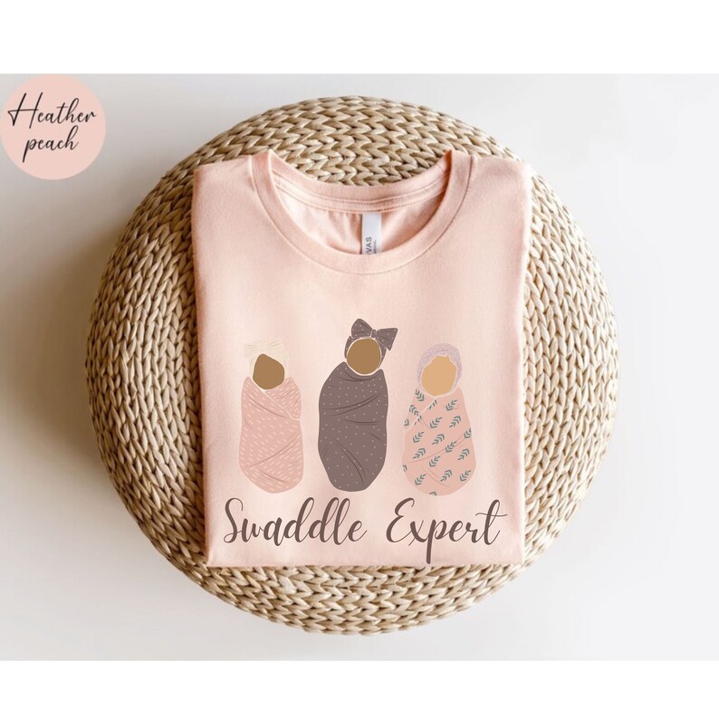 May include: A pink t-shirt with the text "Swaddle Expert" and an illustration of three babies swaddled in different patterns.