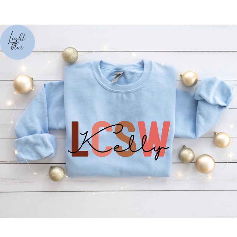 K&ouml;nnte beinhalten: Hellblauer Pullover mit dem Text "LCSW Kelly" in braunen, rosa und orangen Buchstaben.