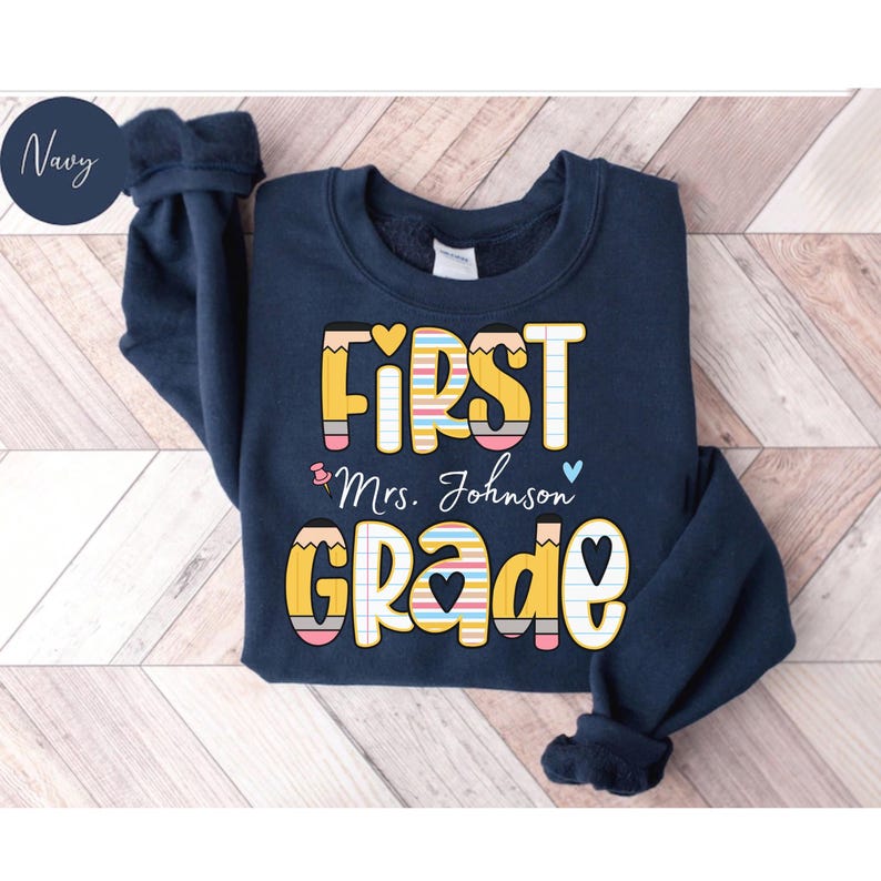 Puede incluir: Sudadera azul marino con las palabras "FIRST Mrs. Johnson GRADE" en un dise&ntilde;o colorido con tem&aacute;tica de l&aacute;pices. El texto es de color amarillo, blanco y colores pastel. La sudadera tiene cuello redondo y mangas largas.