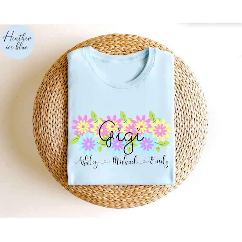 May include: Light blue t-shirt with a floral design and the text "Gigi" in pink and yellow. Below the floral design is the text "Ashley - Michael - Emily" in black.