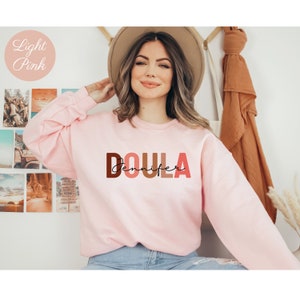 Pu&ograve; includere: Una felpa rosa chiaro con la parola "DOULA" in lettere maiuscole colorate e il nome "Jennifer" scritto in corsivo sotto.