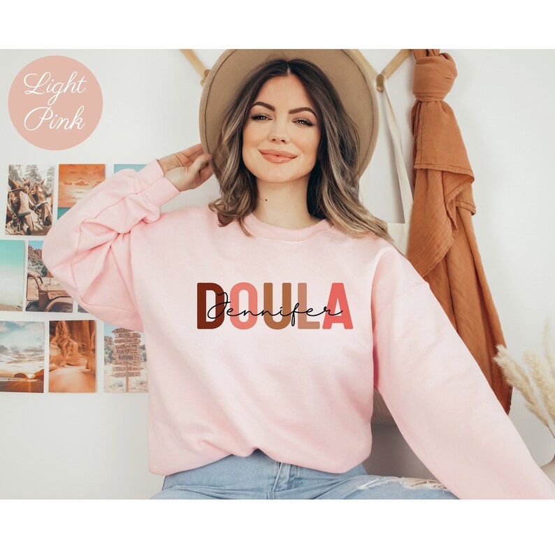 Pu&ograve; includere: Una felpa rosa chiaro con la parola "DOULA" in lettere maiuscole colorate e il nome "Jennifer" scritto in corsivo sotto.