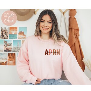 May include: A light pink sweatshirt with the text 'APRN' in brown, orange, and pink letters. The text 'Jennifer' is written in black script below the letters.