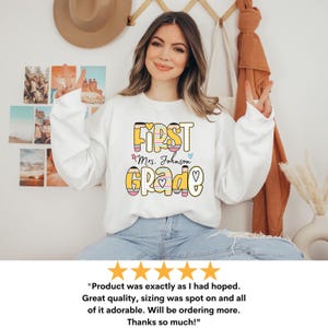 Puede incluir: Sudadera blanca con las palabras "FIRST GRADE" en un dise&ntilde;o colorido de l&aacute;pices. Tambi&eacute;n se incluye el texto "Mrs. Johnson". La sudadera la lleva una persona con vaqueros azules.