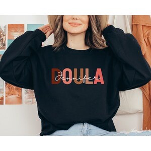 Pu&ograve; includere: Felpa nera con la parola "DOULA" in lettere maiuscole colorate e il nome "Jennifer" scritto in corsivo bianco sotto.