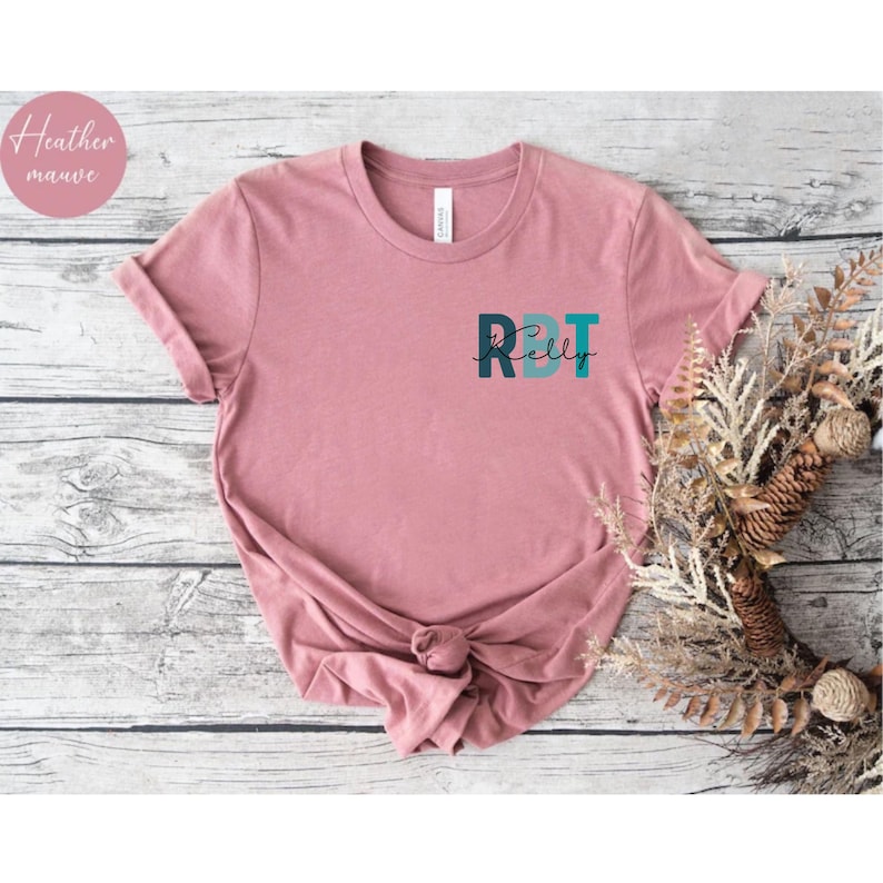 May include: A heather mauve t-shirt with the text "RDT Kelly" in teal and navy blue. The shirt is knotted at the bottom and has rolled-up sleeves. A small circle with the words "Heather mauve" is in the upper left corner.