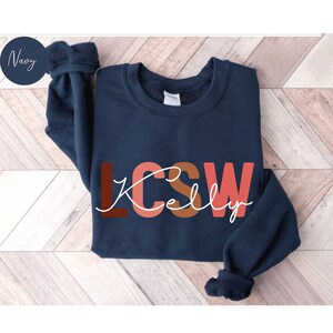 K&ouml;nnte beinhalten: Navyblauer Sweatshirt mit dem Text "LCSW Kelly" in bunten Buchstaben. Die Buchstaben sind in Rot-, Orange- und Rosat&ouml;nen gehalten.