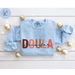 Pu&ograve; includere: Felpa blu chiaro con la parola "DOULA" in lettere colorate e il nome "Jennifer" scritto in corsivo nero sotto.