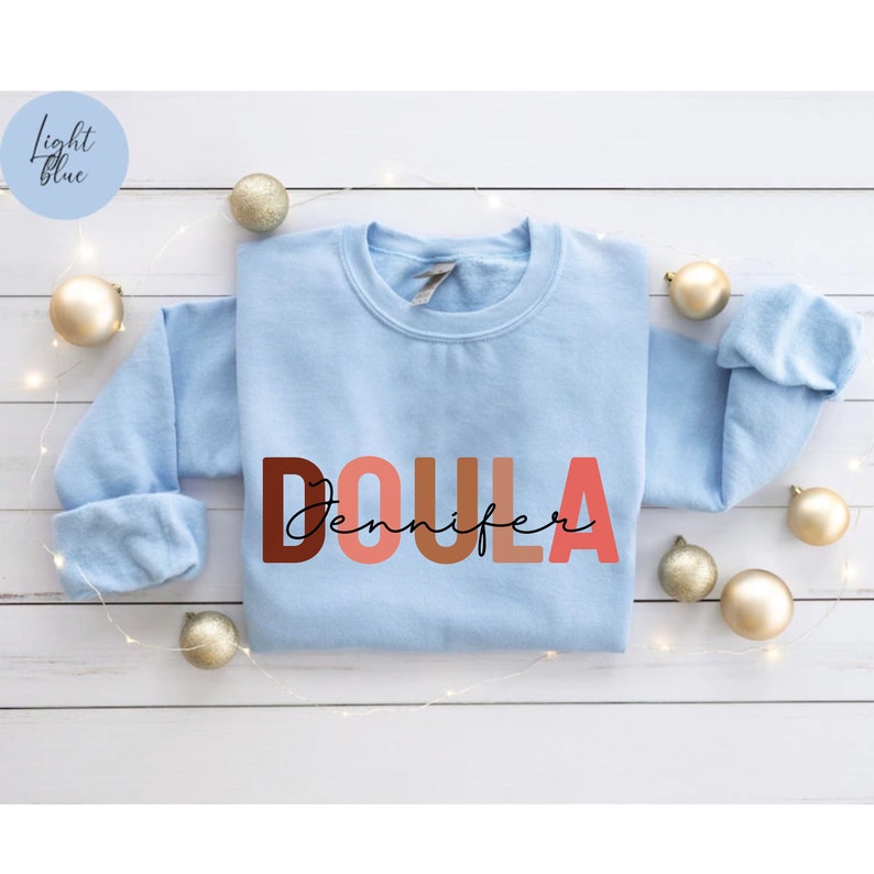 Pu&ograve; includere: Felpa blu chiaro con la parola "DOULA" in lettere colorate e il nome "Jennifer" scritto in corsivo nero sotto.