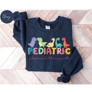 May include: A navy blue sweatshirt with a colorful dinosaur design and the text 'PEDIATRIC physical therapist'.