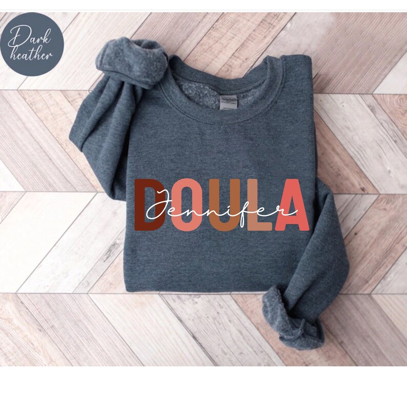 Pu&ograve; includere: Felpa grigia melange scuro con la parola "DOULA" in lettere maiuscole colorate e il nome "Jennifer" scritto in corsivo sotto.
