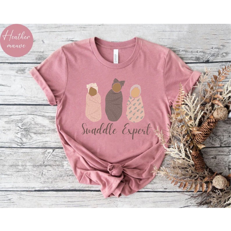 May include: A heather mauve t-shirt featuring a graphic of three swaddled babies and the text "Swaddle Expert".