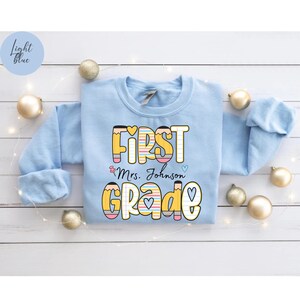 Puede incluir: Sudadera azul claro con las palabras "FIRST GRADE" en un dise&ntilde;o colorido con tem&aacute;tica de l&aacute;pices. El texto "Mrs. Johnson" est&aacute; debajo de la palabra "FIRST". La sudadera est&aacute; decorada con corazones y l&aacute;pices.