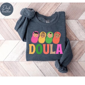 May include: Dark heather grey sweatshirt with a graphic of four babies wrapped in blankets in different colors. The word "DOULA" is written in large letters below the babies, with each letter in a different color.