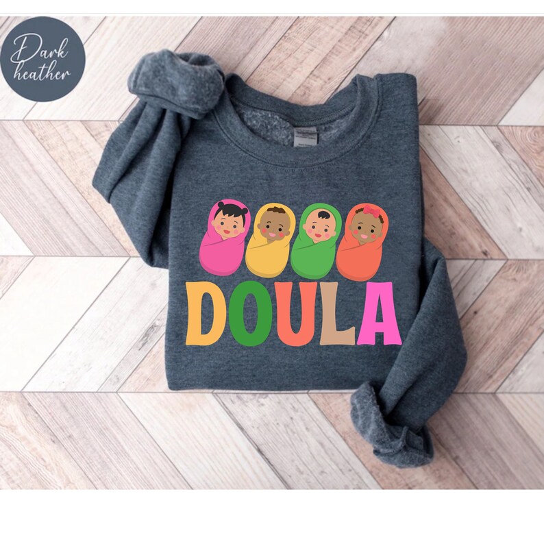 May include: Dark heather grey sweatshirt with a graphic of four babies wrapped in blankets in different colors. The word "DOULA" is written in large letters below the babies, with each letter in a different color.
