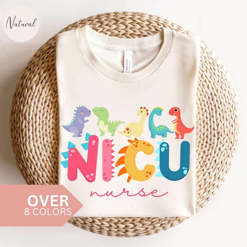 Pu&ograve; includere: T-shirt color crema con dinosauri dei cartoni animati colorati sopra la scritta "NICU nurse". Le lettere sono rosa, arancioni e blu. La maglietta ha anche la scritta "OVER 8 COLORS" in un riquadro rosa.