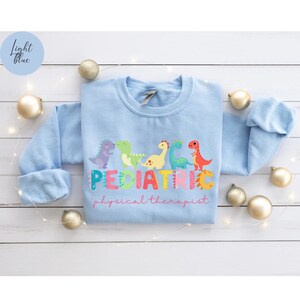 May include: Light blue sweatshirt with a graphic of colorful dinosaurs spelling out 'PEDIATRIC' and the words 'physical therapist' below.
