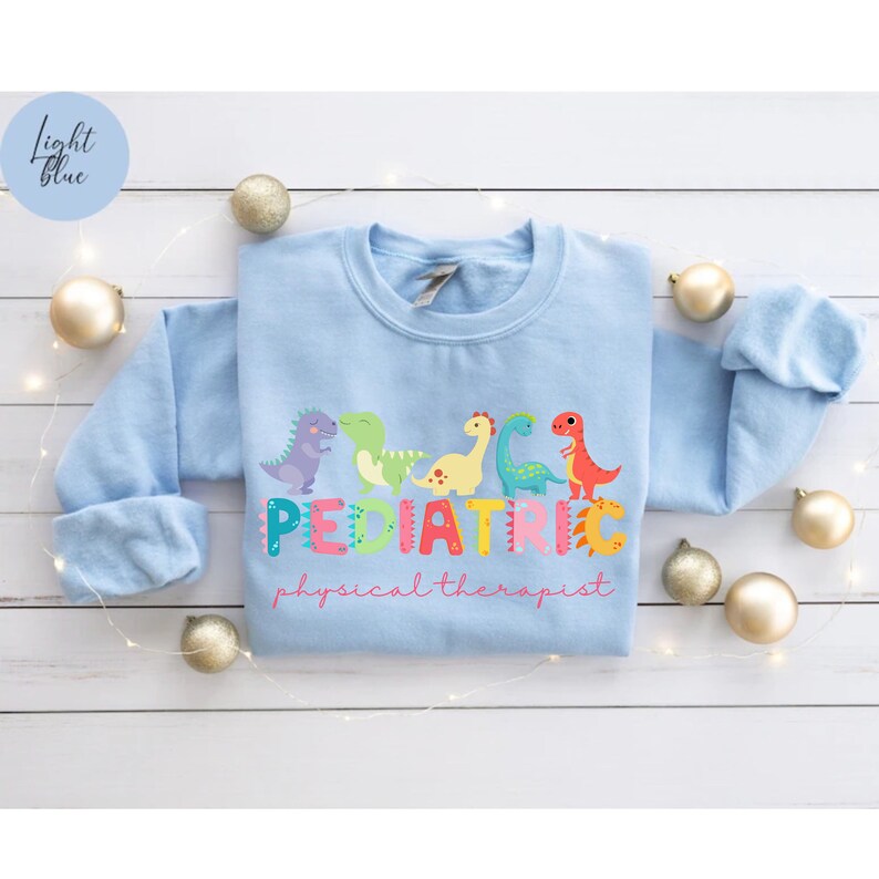 May include: Light blue sweatshirt with a graphic of colorful dinosaurs spelling out 'PEDIATRIC' and the words 'physical therapist' below.
