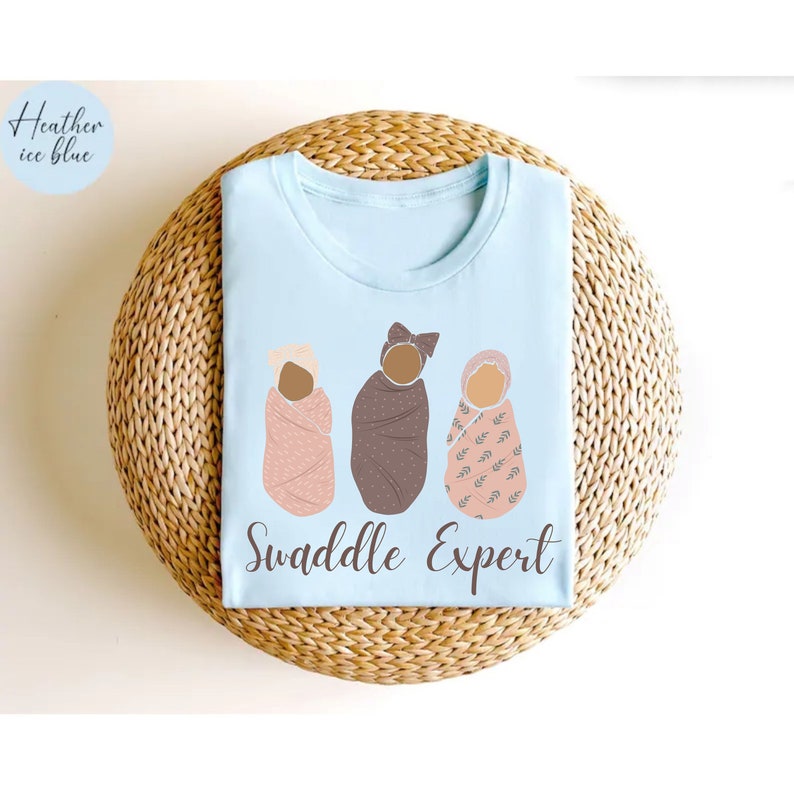 May include: A light blue t-shirt with the text "Swaddle Expert" and a graphic of three babies swaddled in different patterned blankets.