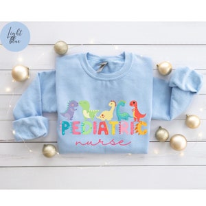May include: Light blue sweatshirt with a graphic of colorful dinosaurs and the text "PEDIATRIC nurse".