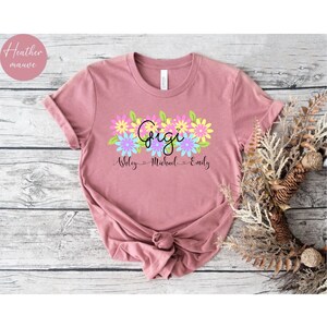 May include: Heather mauve t-shirt with a floral design and the word "Gigi" in pink, yellow, and blue. Below the word "Gigi" are the names "Ashley", "Michael", and "Emily" in black.