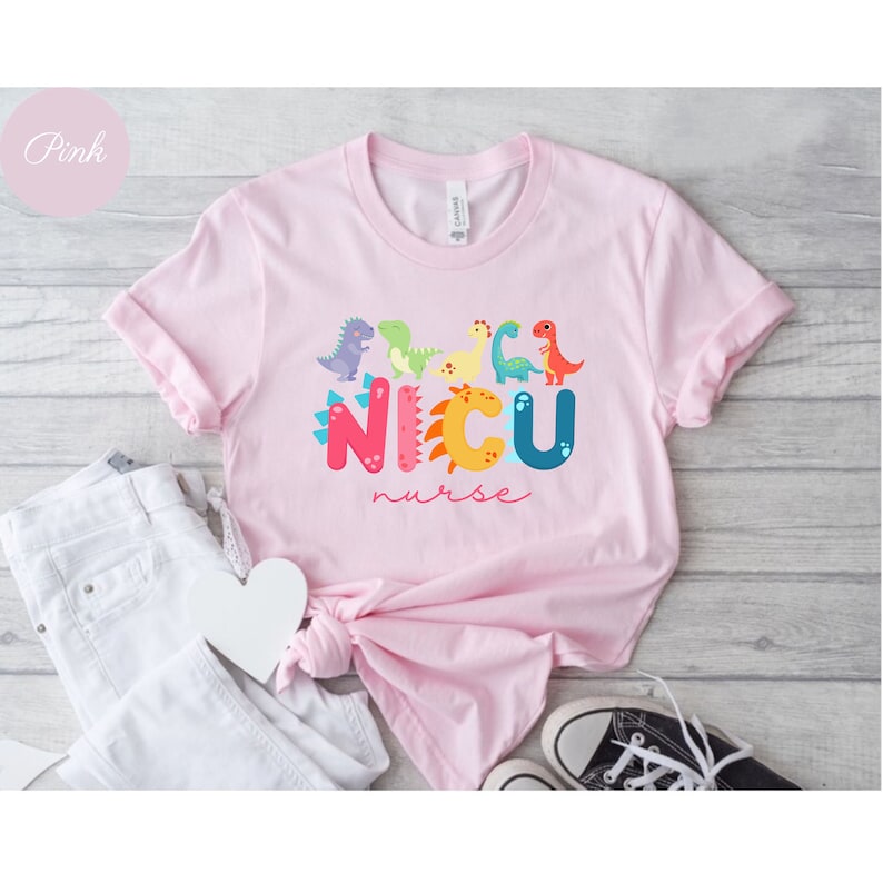 Pu&ograve; includere: T-shirt rosa chiaro con la scritta "NICU nurse" in lettere colorate e una fila di dinosauri dei cartoni animati. La maglietta &egrave; annodata, con pantaloni bianchi e scarpe da ginnastica bianche e nere sullo sfondo.