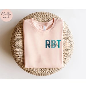 May include: A light peach-colored t-shirt with the letters "RBT" in teal and navy blue, with the name "Kelly" in a script font underneath. The shirt is displayed on a woven, circular surface. The words "Heather peach" are in a circle in the upper left corner.
