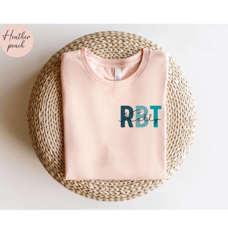 May include: A light peach-colored t-shirt with the letters "RBT" in teal and navy blue, with the name "Kelly" in a script font underneath. The shirt is displayed on a woven, circular surface. The words "Heather peach" are in a circle in the upper left corner.